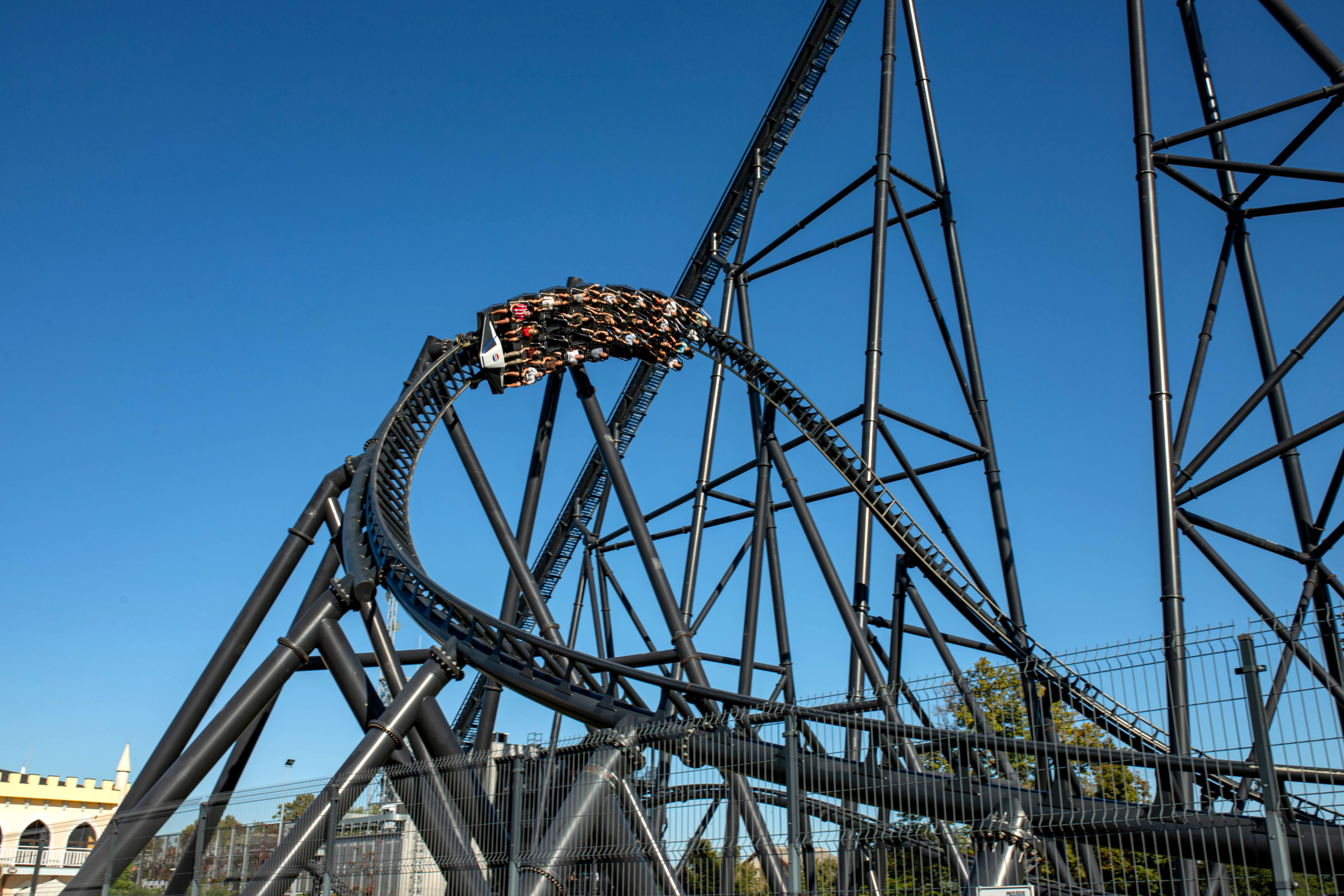 High-intensity coaster turn Zator Black steel roller coaster curve with a packed train riding through the track under a clear blue sky.