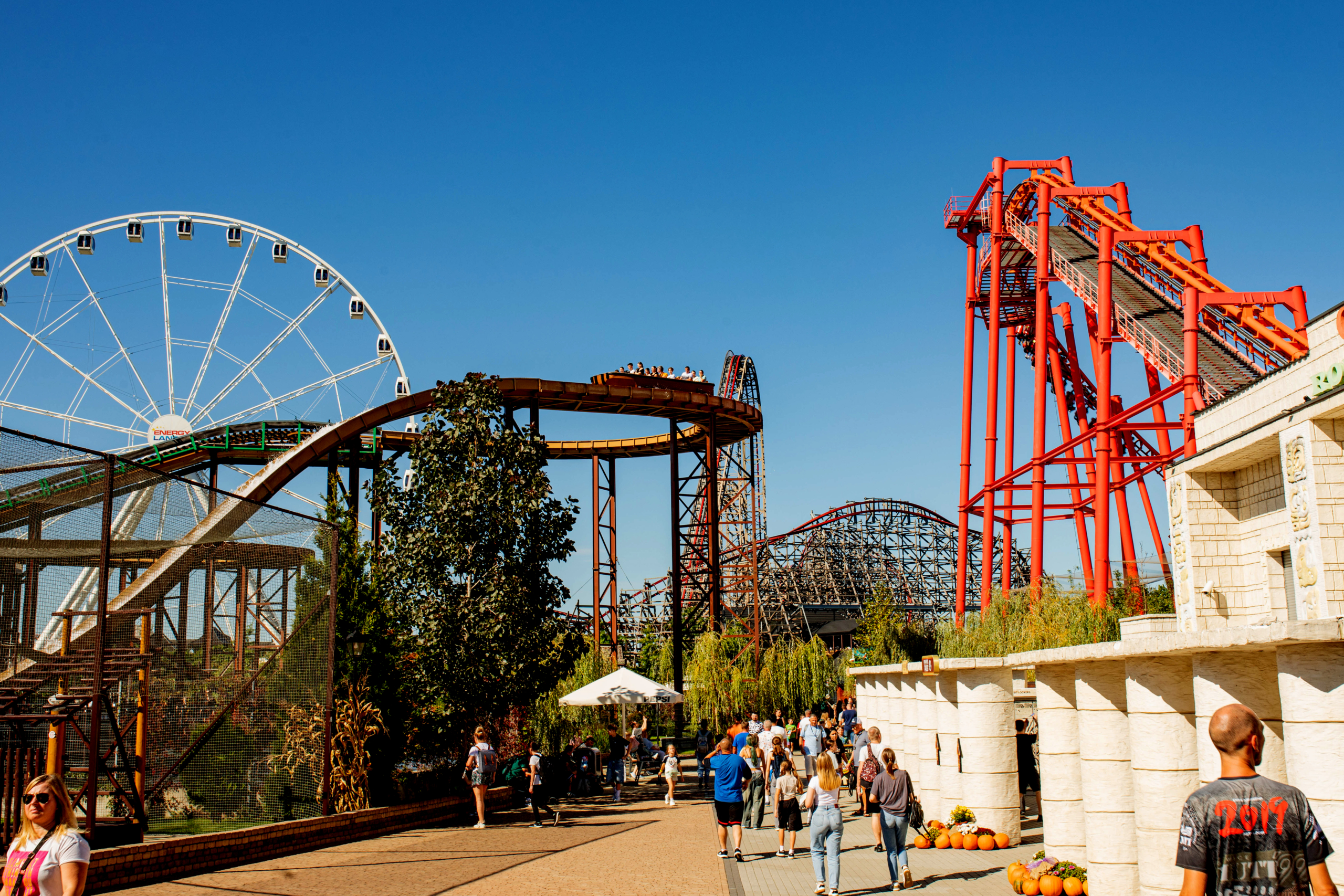 Zator theme park panorama of top attractions Wide view of Energylandia with the Ferris wheel and red steel coaster structures, visitors walking along the central path.